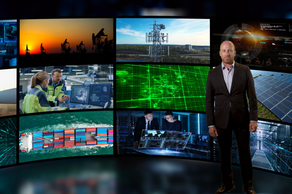 Video still of a spokesman in a suit in front of a virtual background of video monitors depicting technology